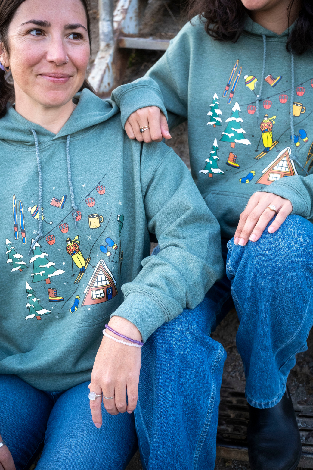 Two people wearing teal hoodies with colorful graphic designs sitting on a wooden bench.