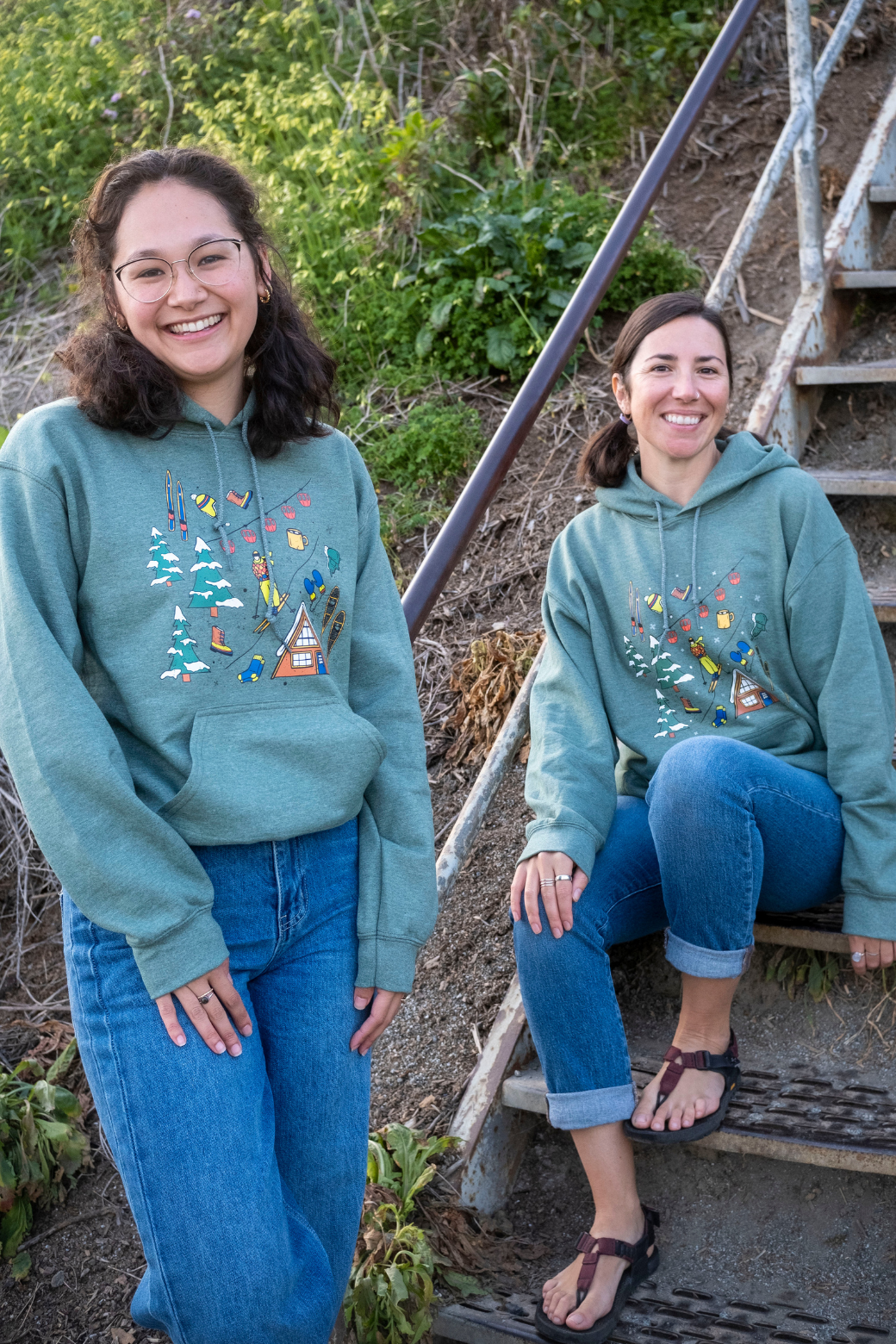 Two women wearing matching teal hoodies with a colorful design outdoors.