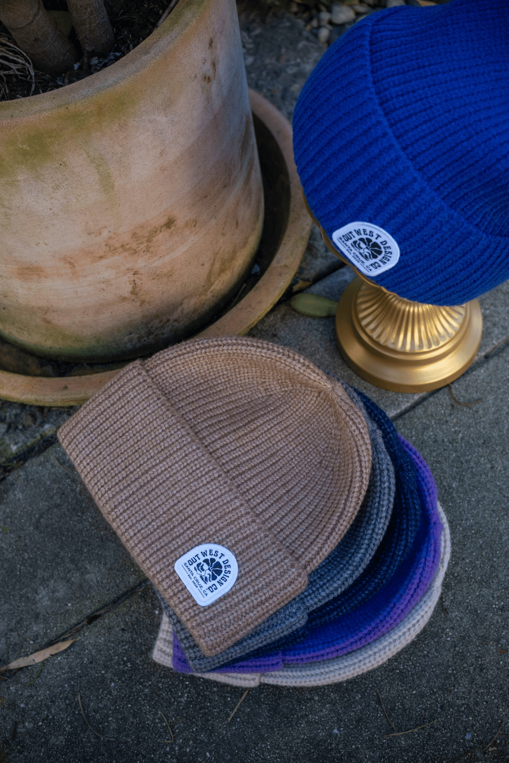 Three beanies in brown, blue, and purple on a stone surface with a plant pot and gold lamp in the background.