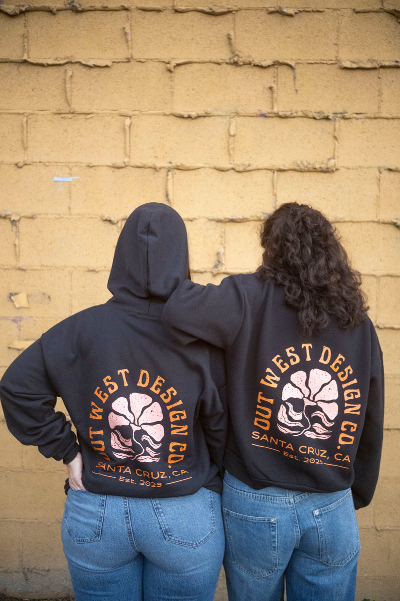 Two people wearing black hoodies with 'Out West Design Co.' branding against a beige brick wall.