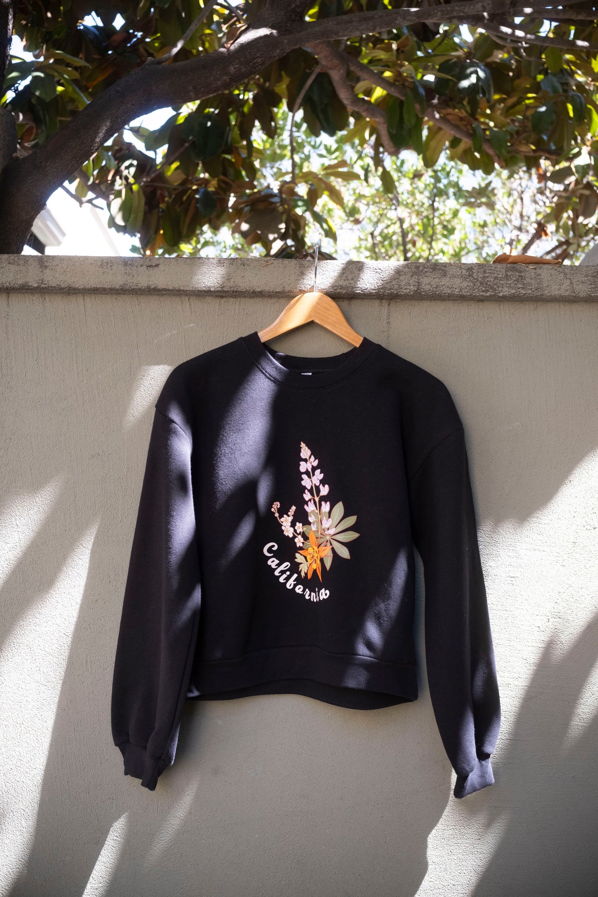 Black sweatshirt with floral design and text on a hanger against a wall with tree shadows.