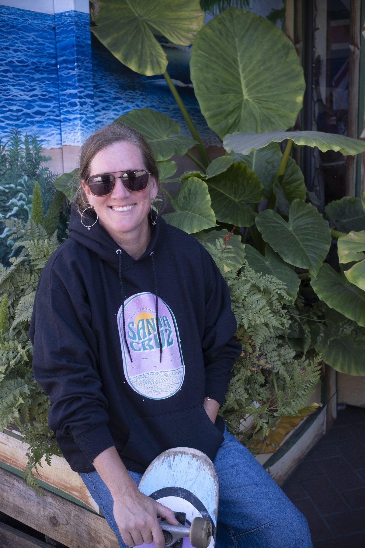 Person wearing a black hoodie with a graphic design, holding a skateboard, sitting among large green leaves.