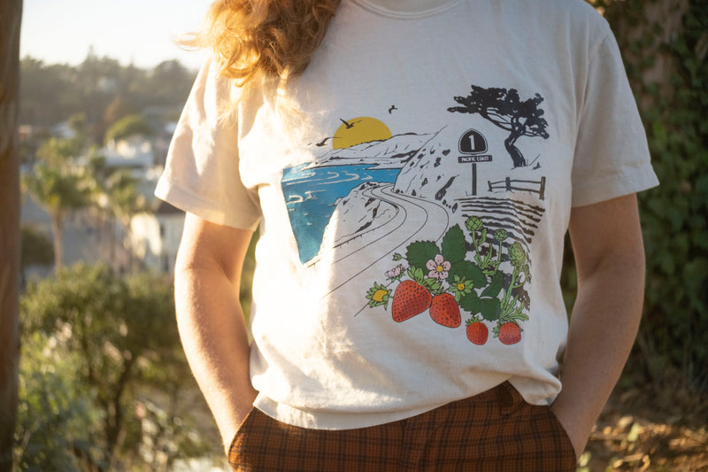 Person wearing a white t-shirt with a scenic and strawberry design, standing outdoors.