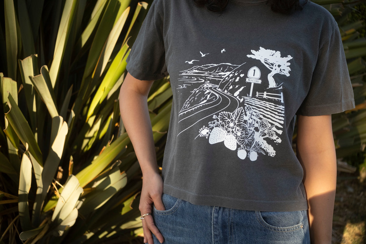 Person wearing a dark gray t-shirt with a white landscape design, standing in front of green plants.