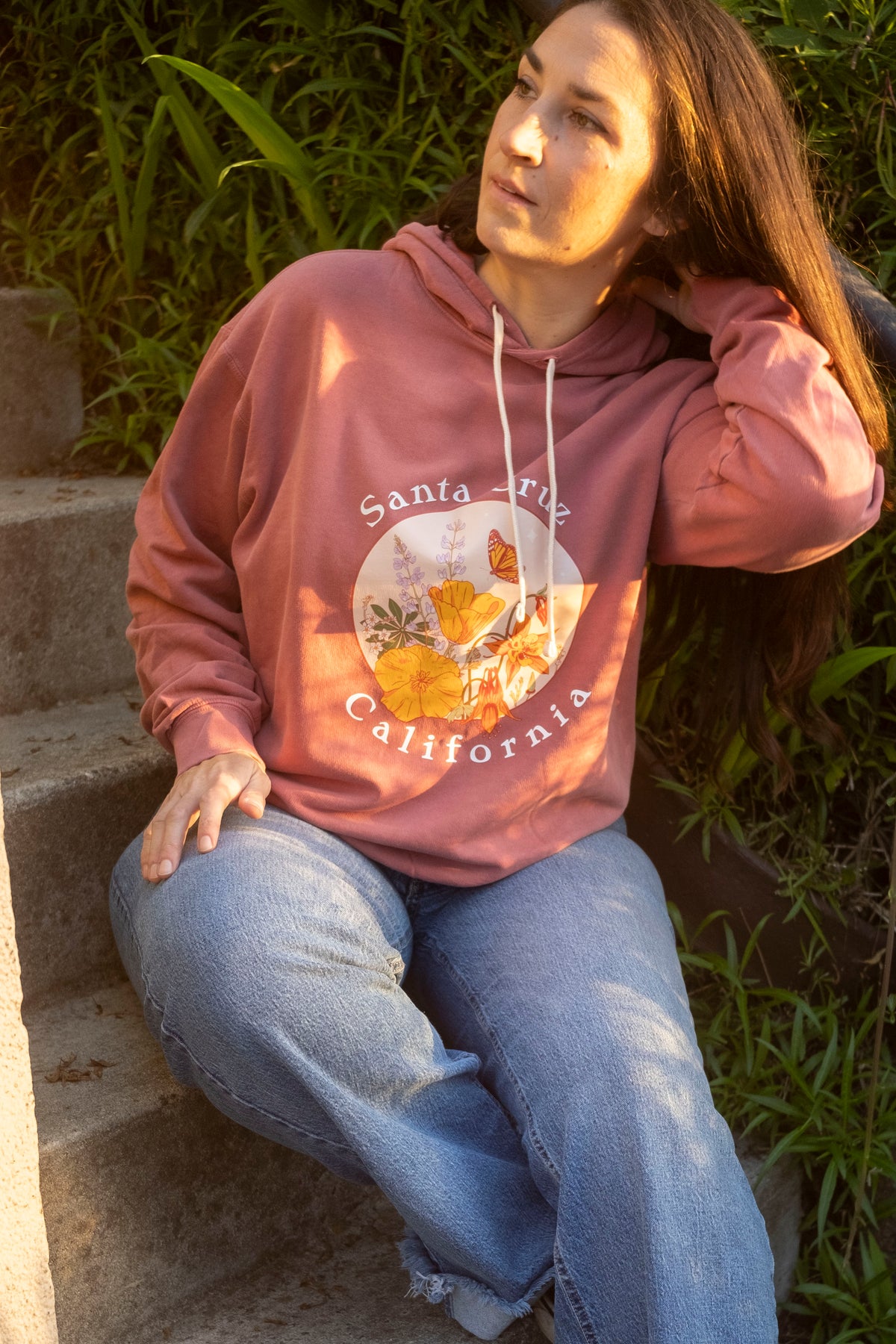 Person wearing a pink hoodie with a 'Santa Cruz California' design, sitting on steps outdoors.