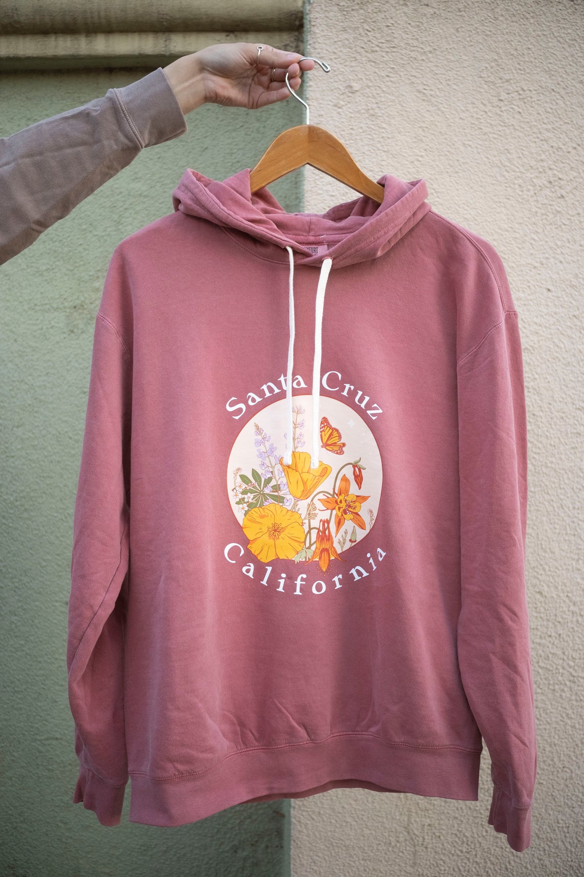 Pink hoodie with 'Santa Cruz California' design on a hanger against a textured wall.