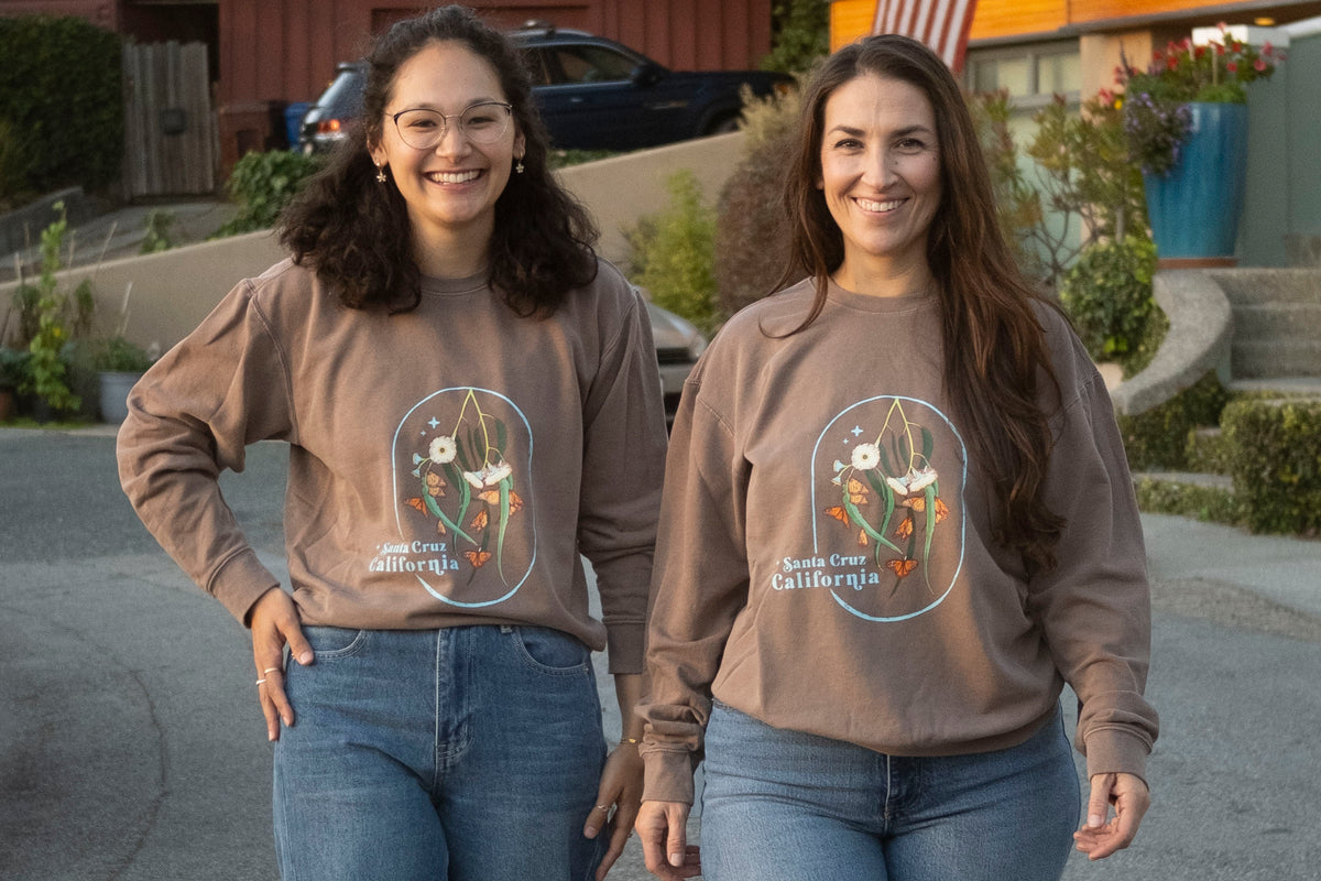 Two women wearing brown sweatshirts with a graphic design outdoors.
