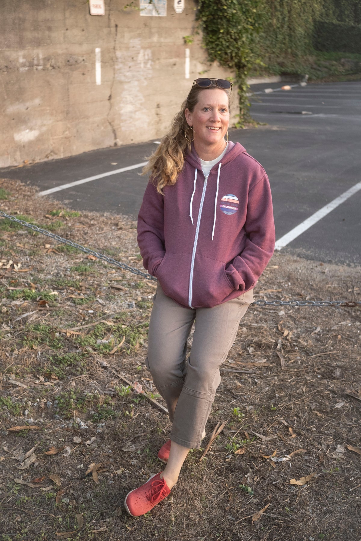 Woman wearing a maroon hoodie with a logo, standing in a parking lot.