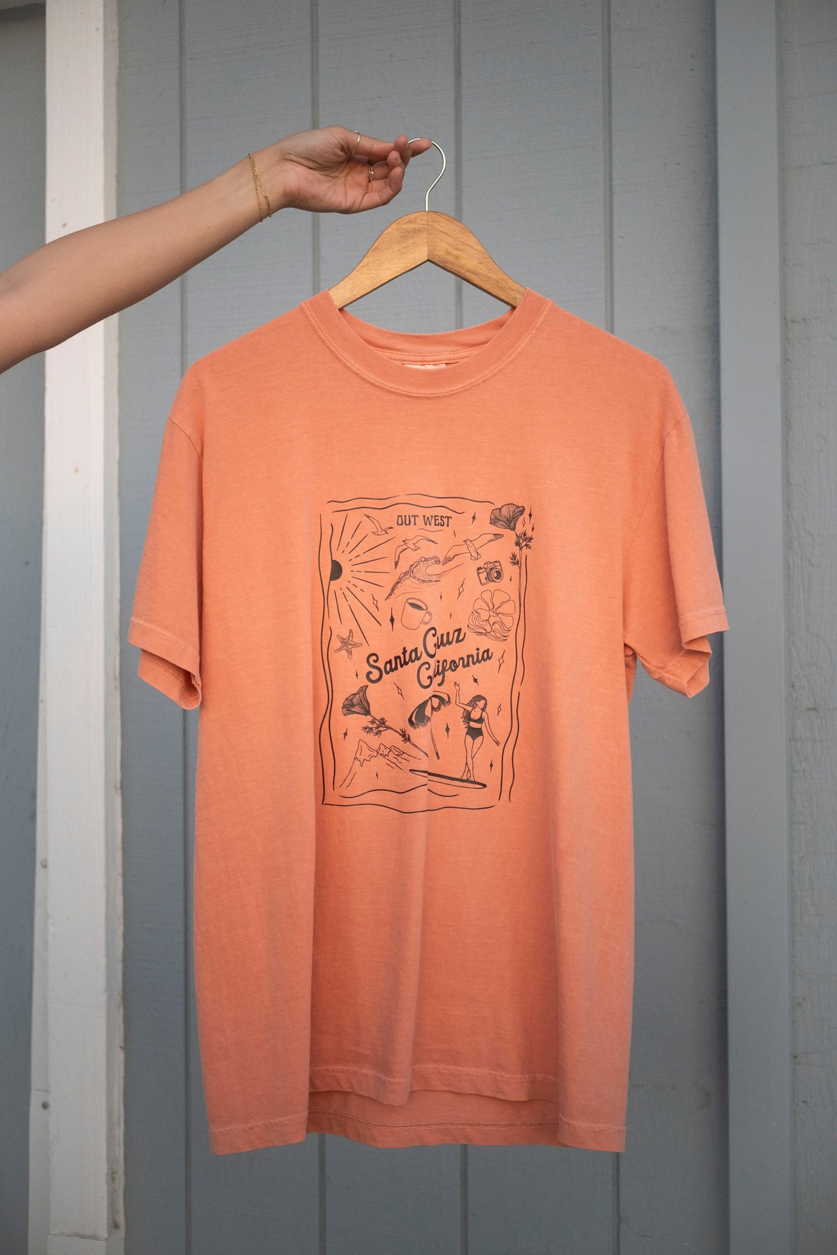 Orange t-shirt with graphic design on a hanger against a gray wooden panel background