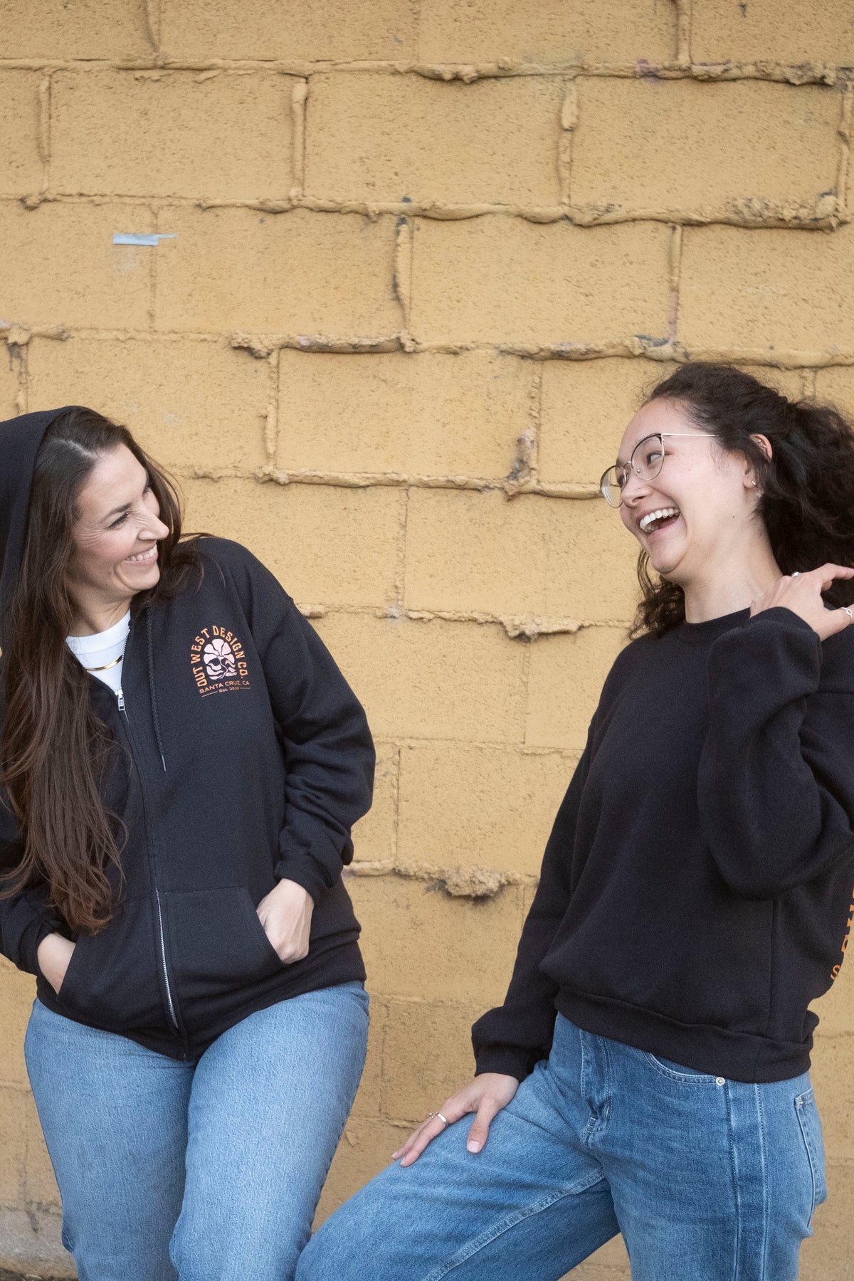 Two women wearing black hoodies in front of a textured beige wall