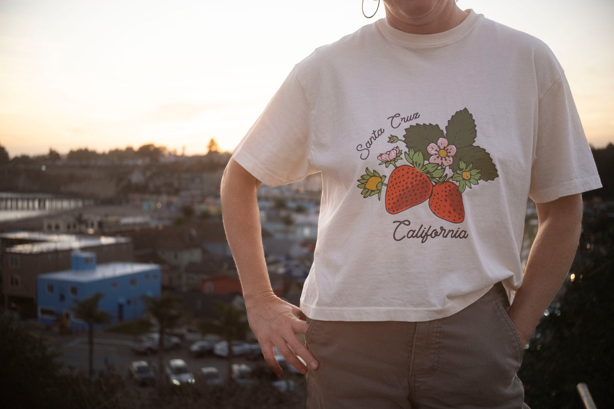 Person wearing a t-shirt with a strawberry design and text, standing against an urban backdrop.