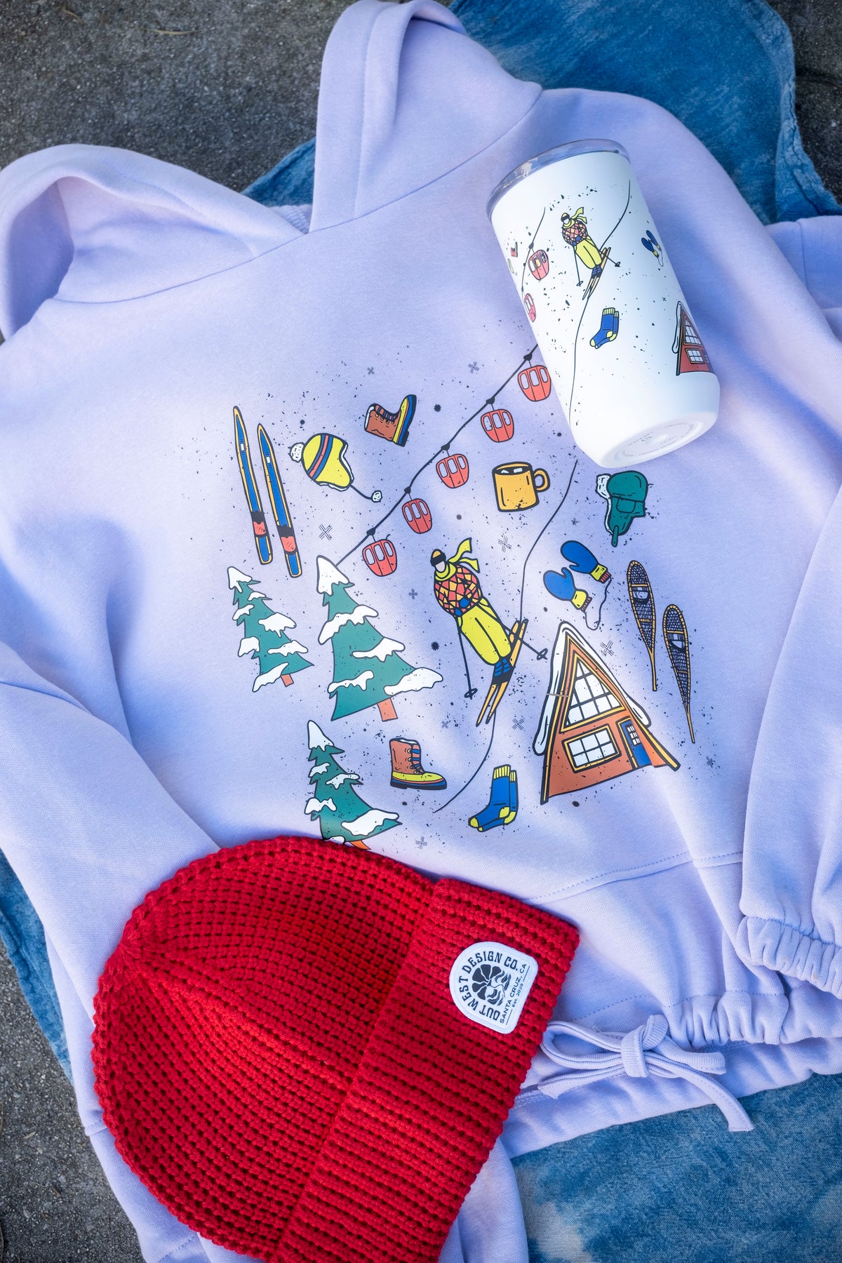 Light purple hoodie with winter-themed design, red beanie, and white mug on a textured surface.