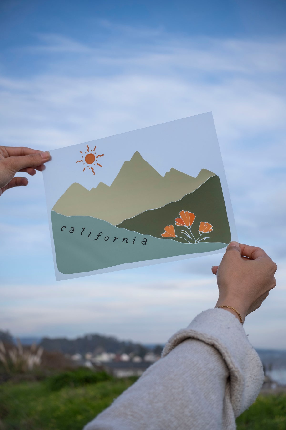 Person holding a print with mountains and sun design against a scenic background