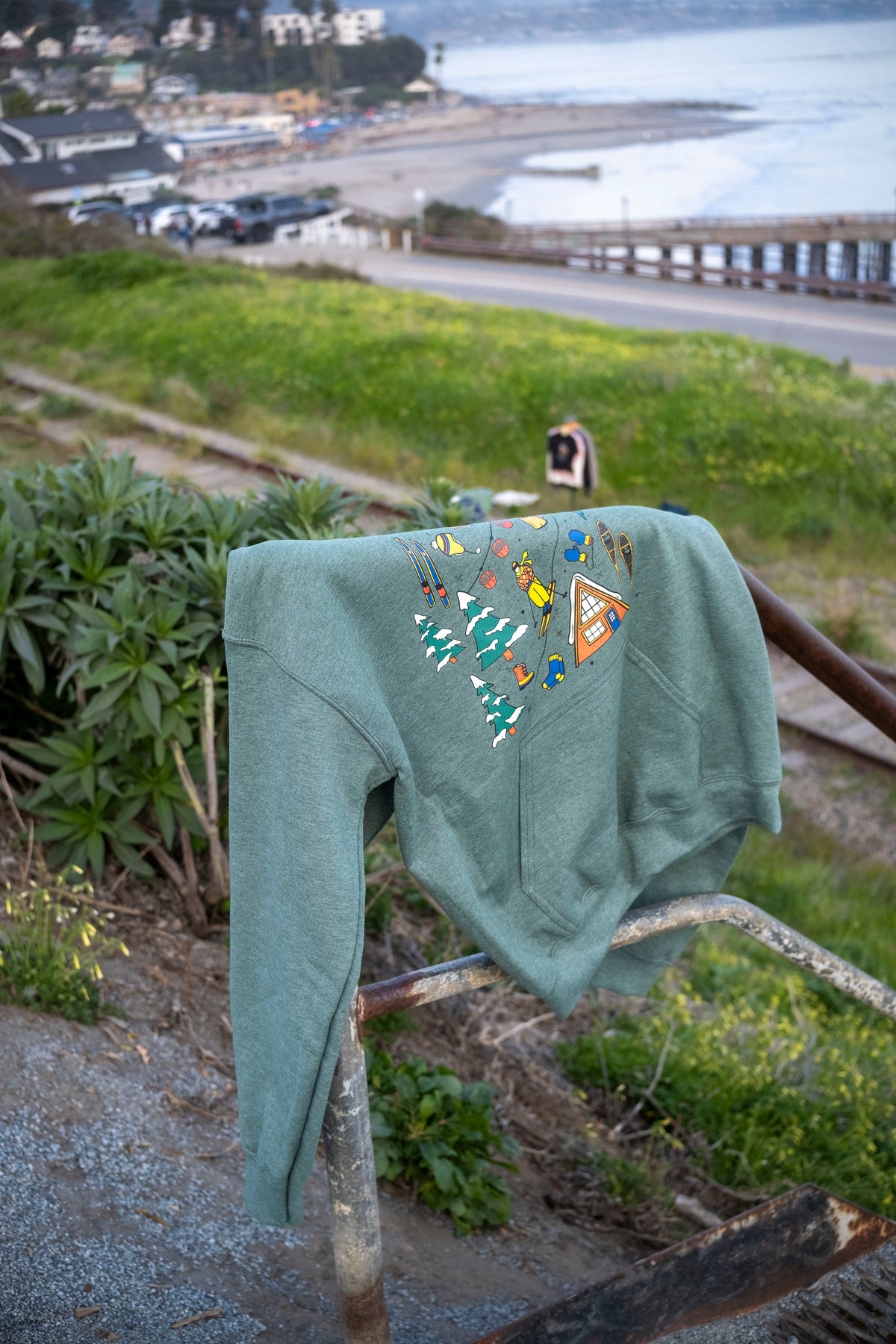 Green hoodie with colorful designs hanging on a metal railing with a scenic background