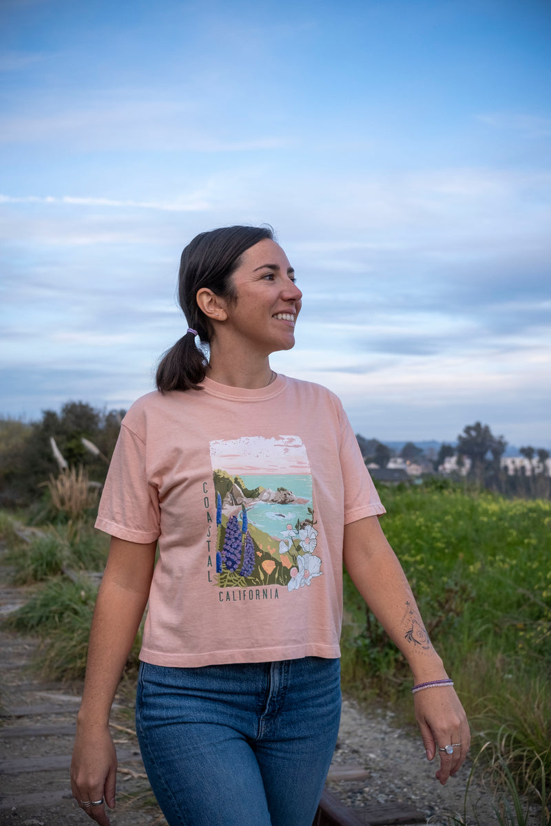 Woman wearing a pink t-shirt with a scenic graphic, standing outdoors with a natural background.