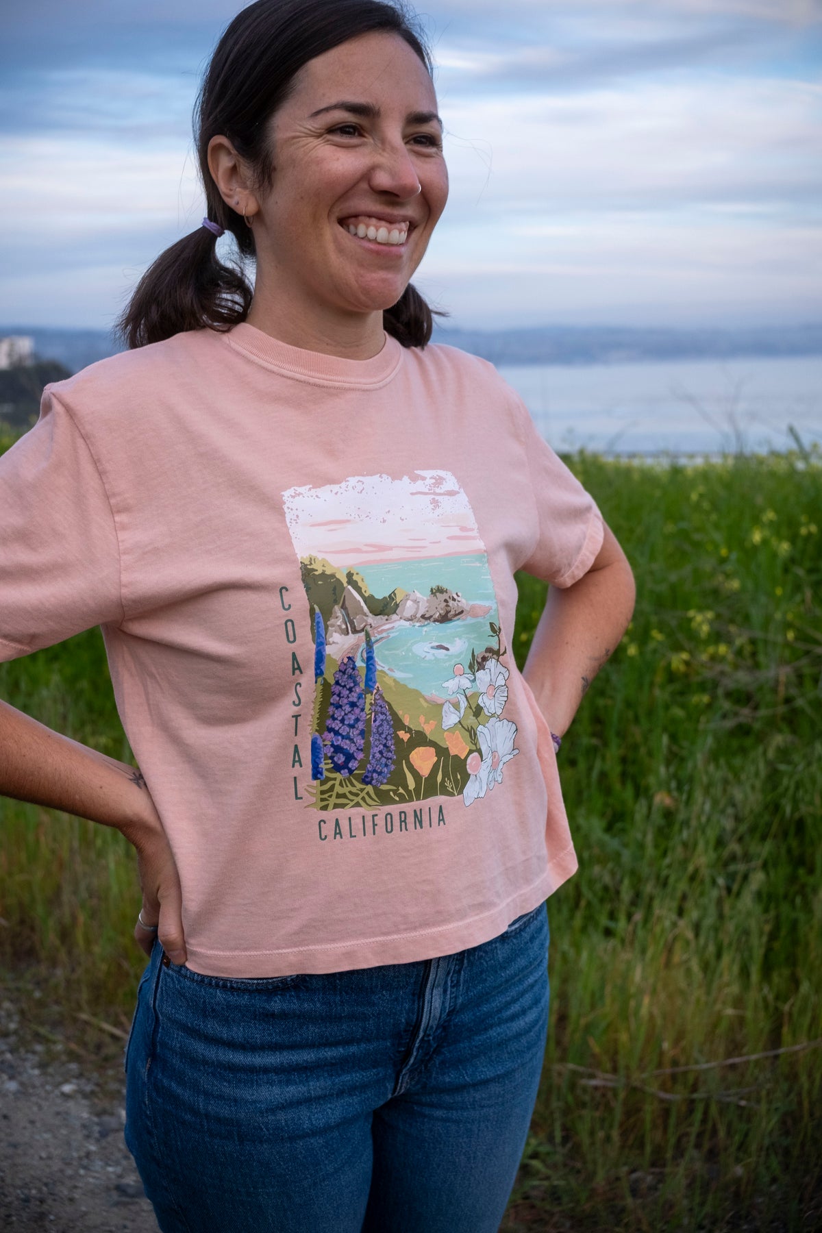 Woman wearing a pink t-shirt with a graphic design of a coastal scene, standing in a field with ocean view.