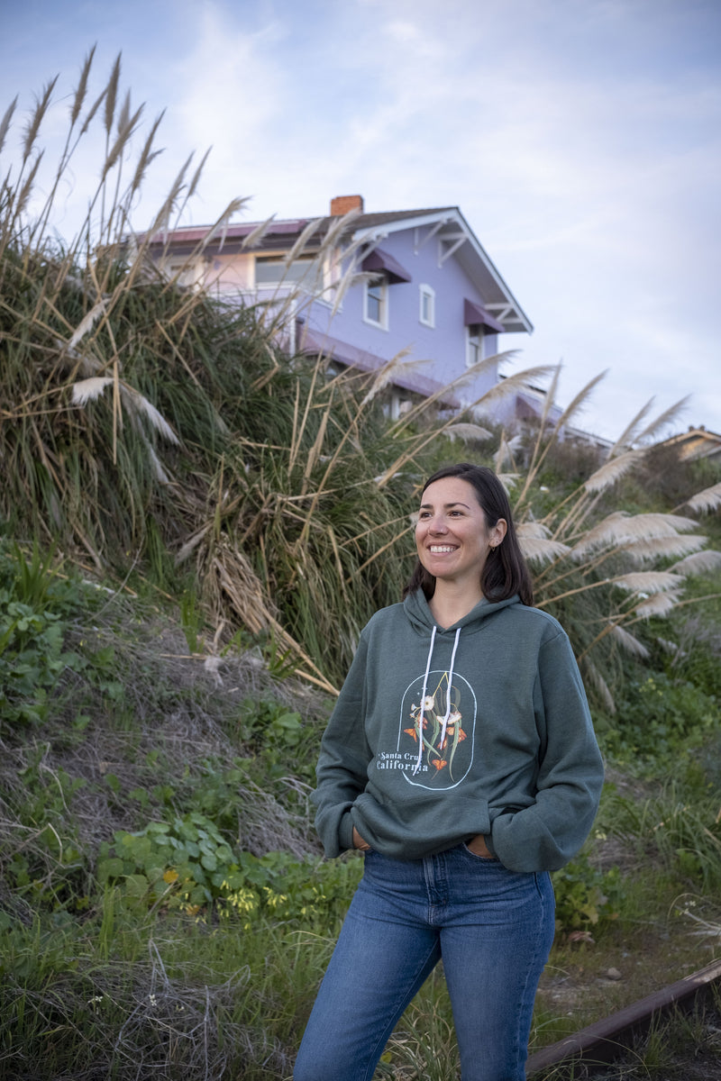 Woman wearing a green hoodie with a graphic design, standing outdoors with tall grass and a house in the background.