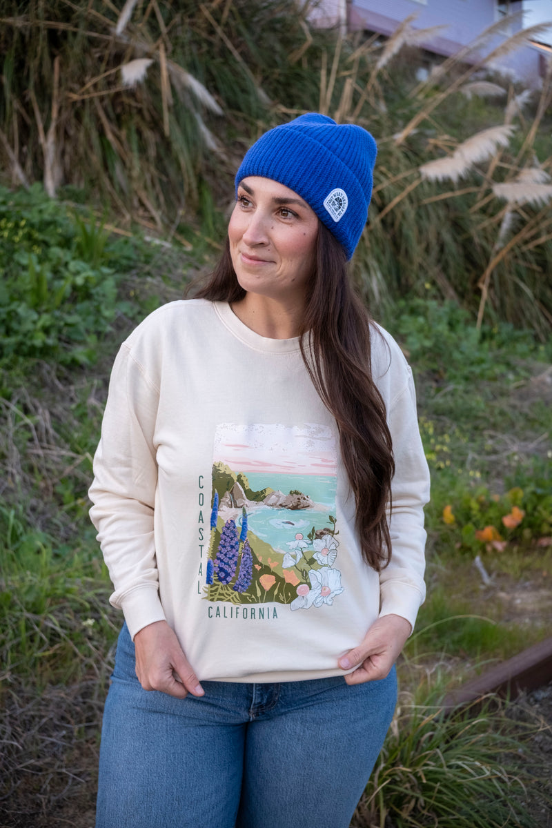 Woman wearing a cream sweatshirt with a scenic print, blue jeans, and a blue beanie outdoors.