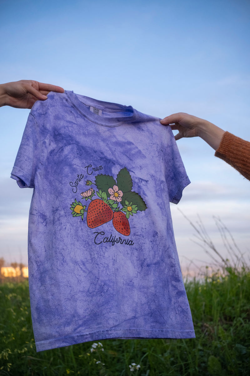 Purple tie-dye t-shirt with strawberry and floral design held against a natural background