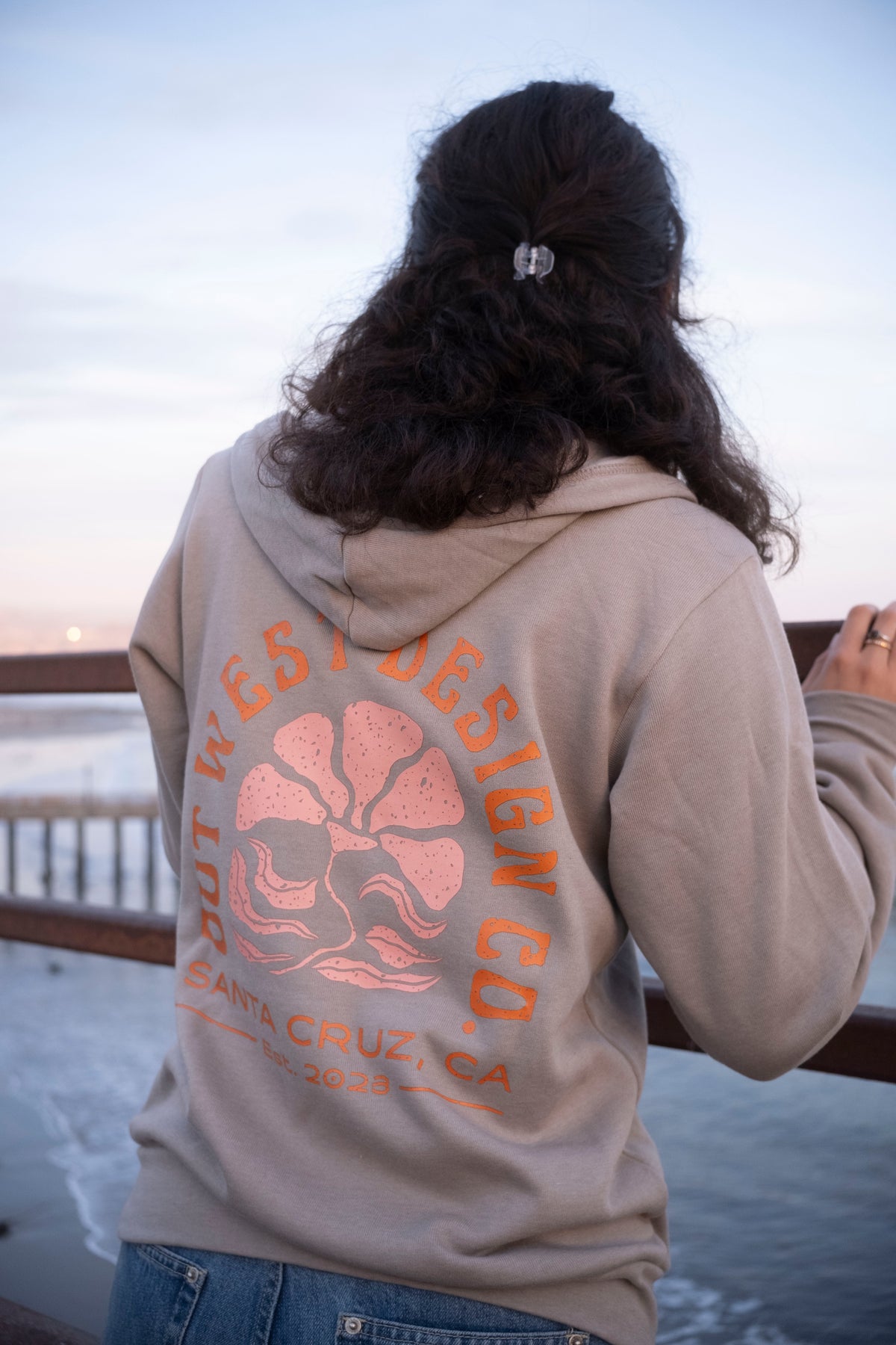 Person wearing a hoodie with a design on the back, standing by a body of water.