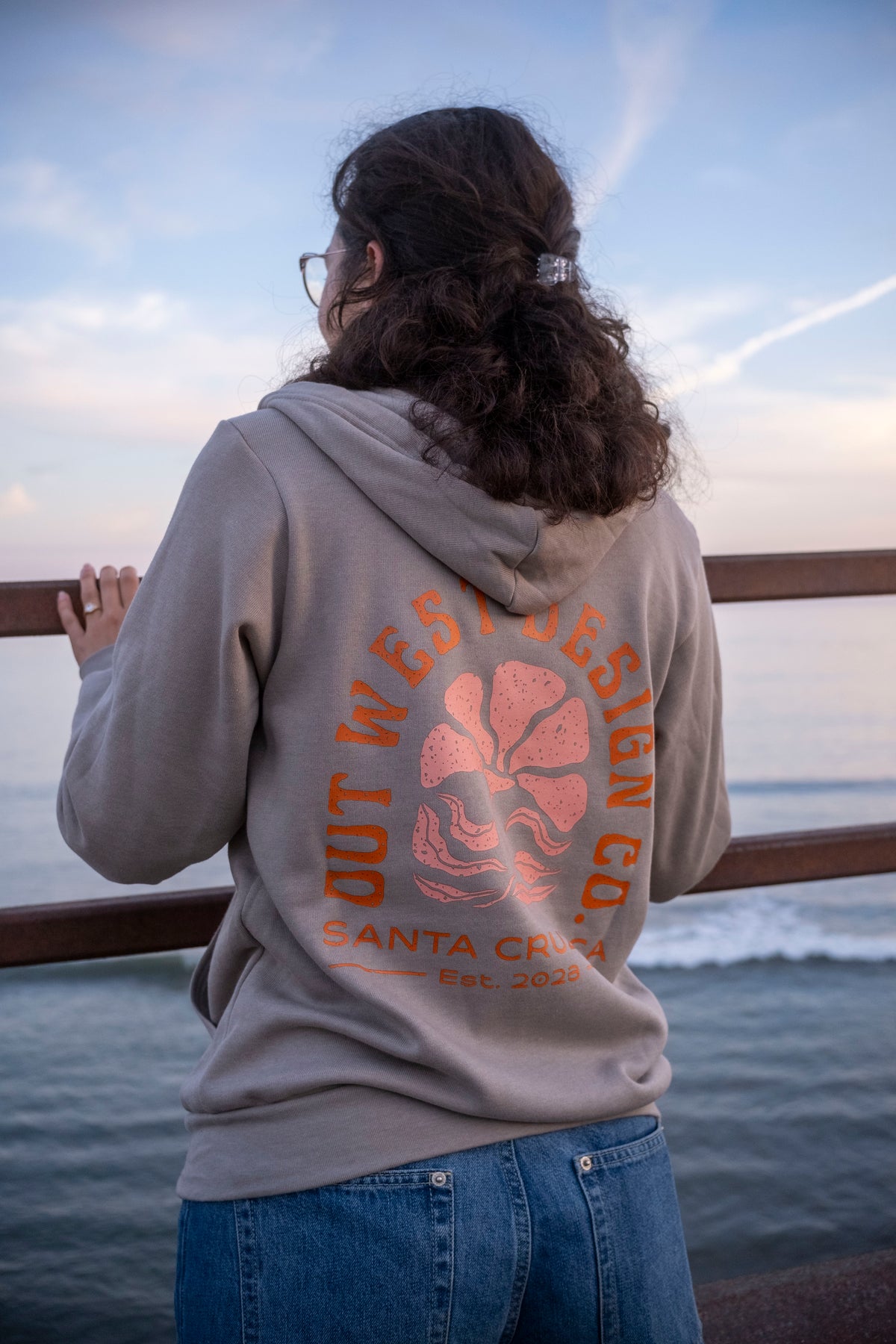 Person wearing a hoodie with a logo and text, standing by a waterfront.