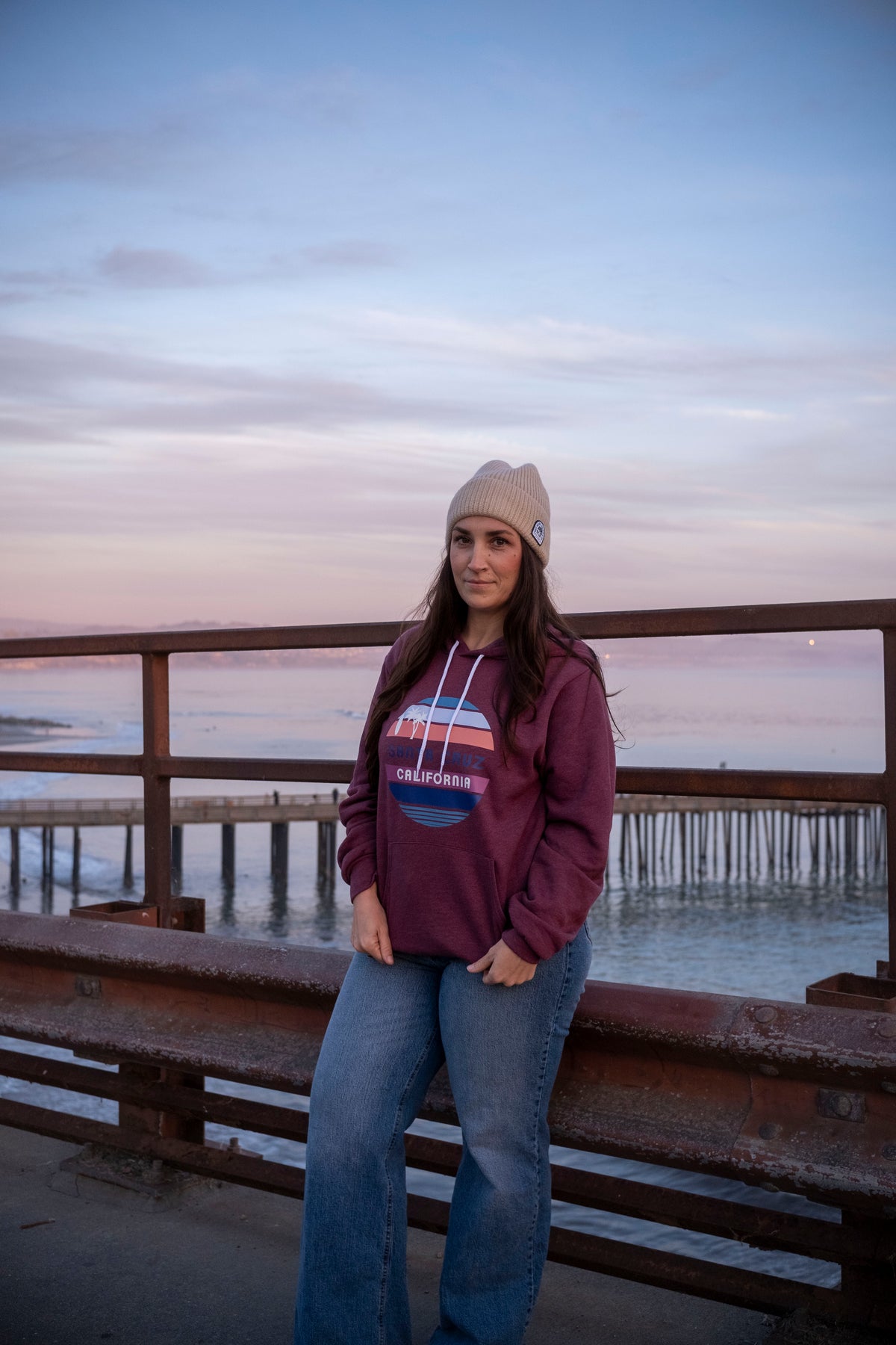 Person wearing a maroon hoodie and beige beanie standing on a pier with water and sky in the background