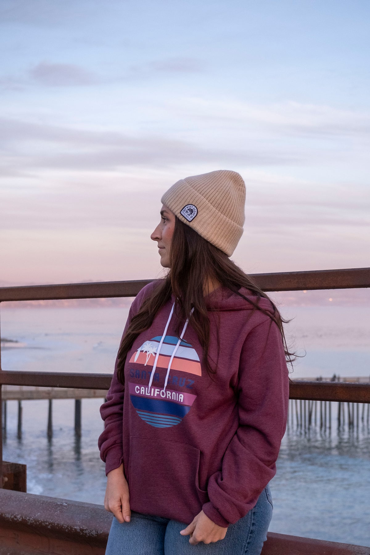 Person wearing a maroon hoodie with a logo and text, standing by a waterfront at sunset.