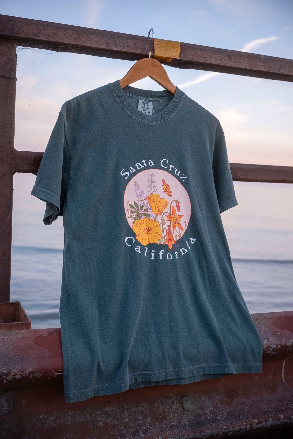 A green Santa Cruz tee hangs from a railing next to the ocean. 