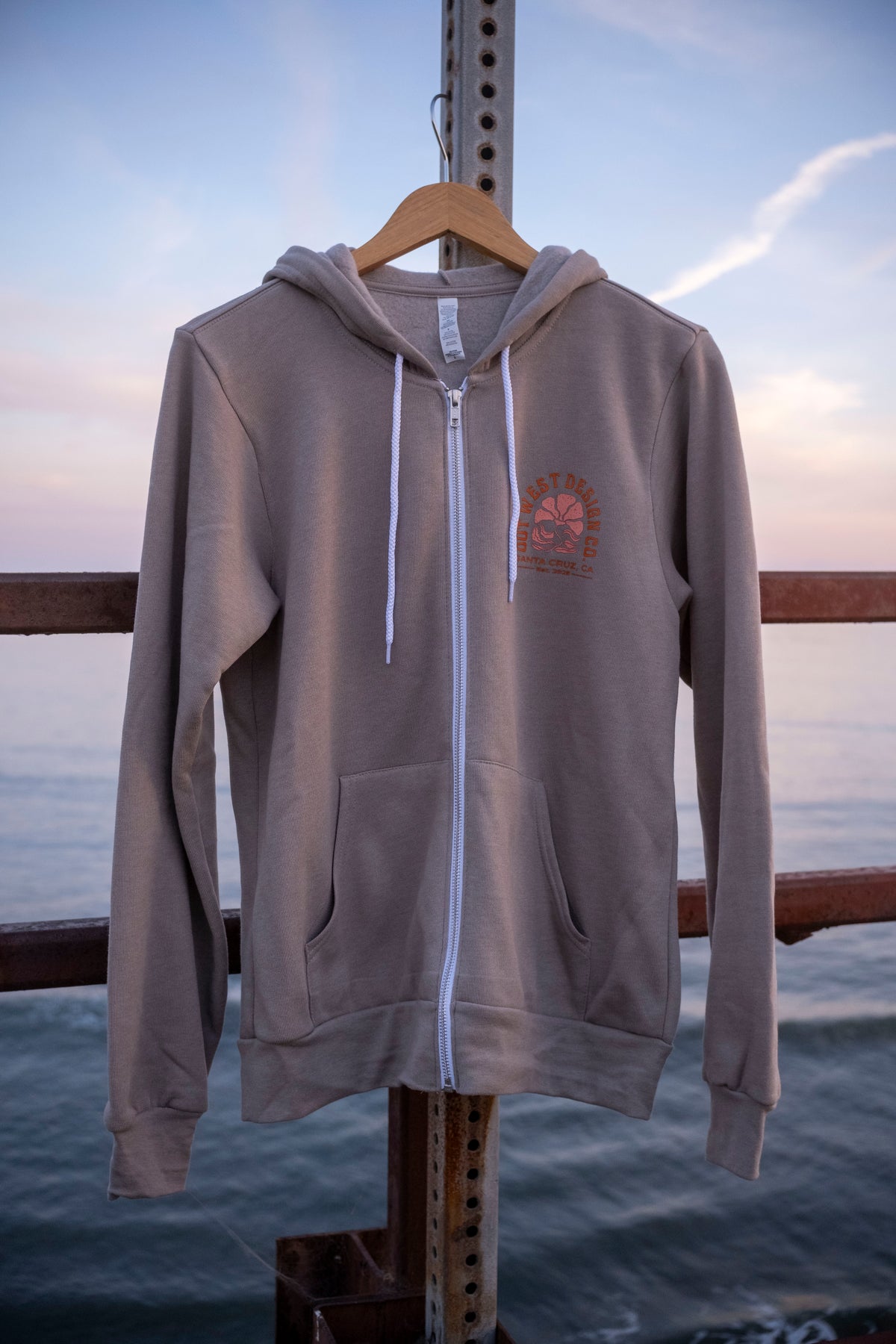 Gray hoodie with a logo on a hanger against a sunset sky.