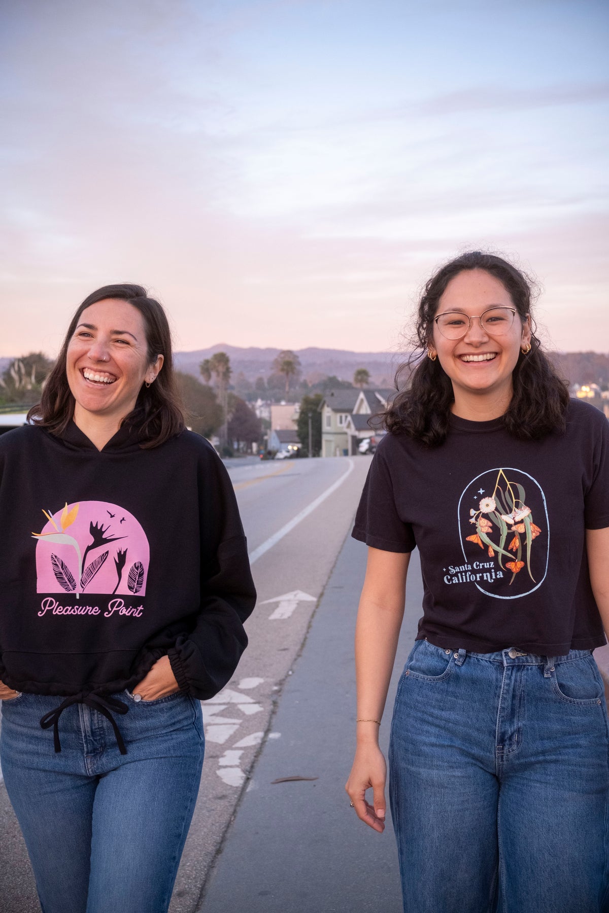 Two women standing on a road, one wearing a black hoodie with a pink design and the other wearing a black t-shirt with a white design.
