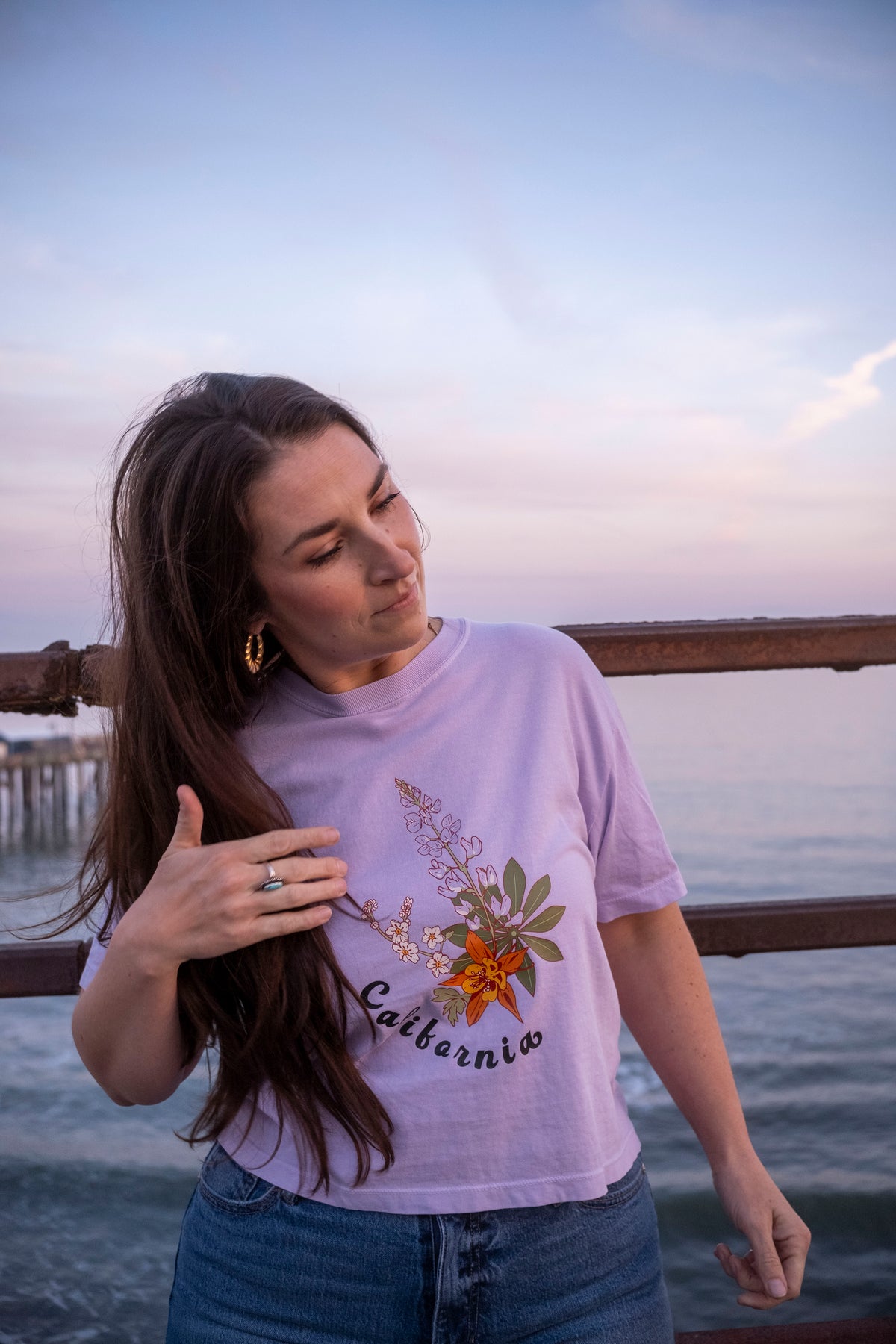 Woman wearing a purple 'California' t-shirt with floral design by the water at sunset.