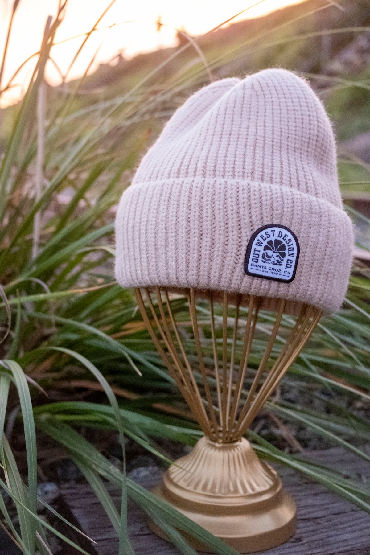 Beige knit beanie with a logo on a wooden stand with grass in the background