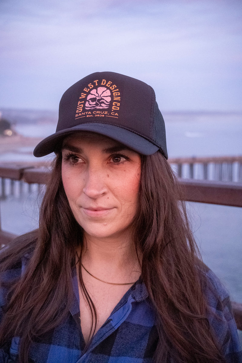Woman wearing a cap with text, standing on a pier with water and sky in the background