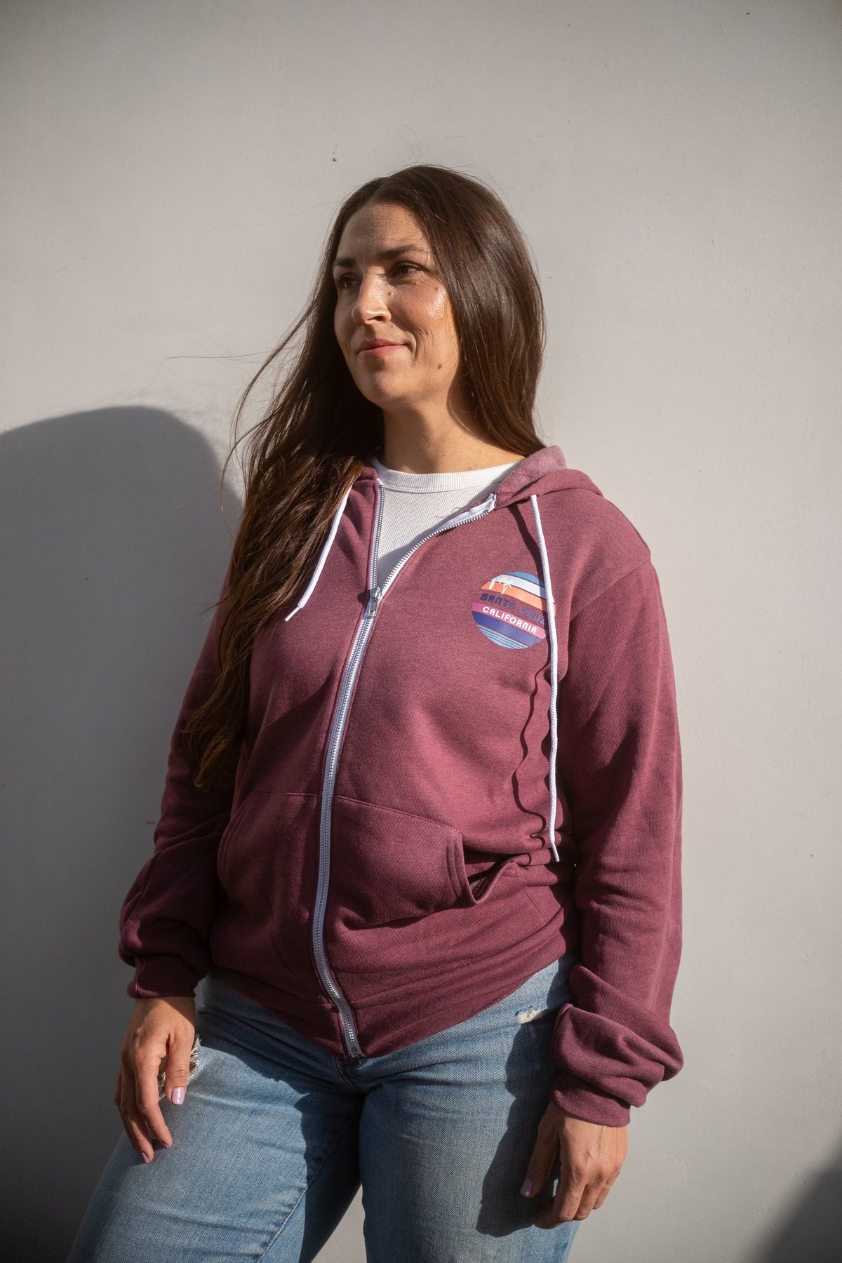 Person wearing a maroon zip-up hoodie with white drawstrings and a circular multicolor 'Santa Cruz, California' chest graphic, paired with blue jeans, standing against a plain light gray wall.