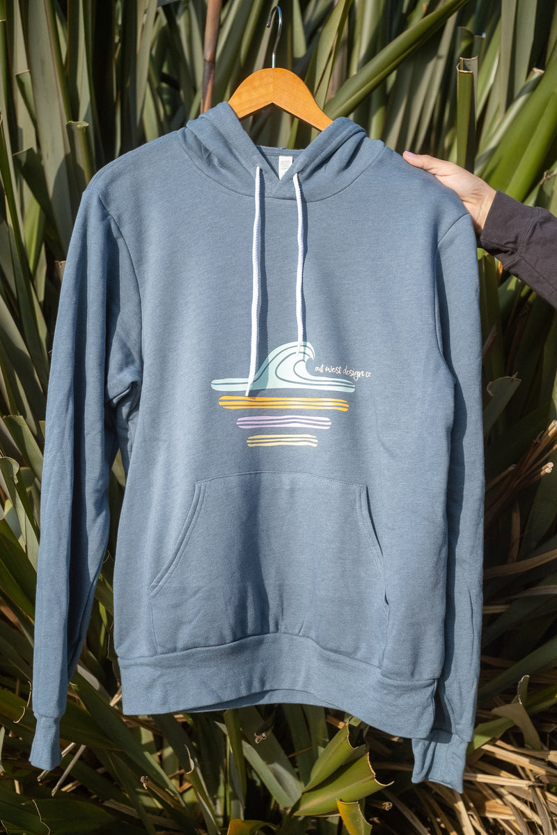 A dusty blue pullover hoodie with a colorful wave graphic on the chest, hanging on a wooden hanger against a background of green tropical leaves. A hand is holding the sleeve of the hoodie.