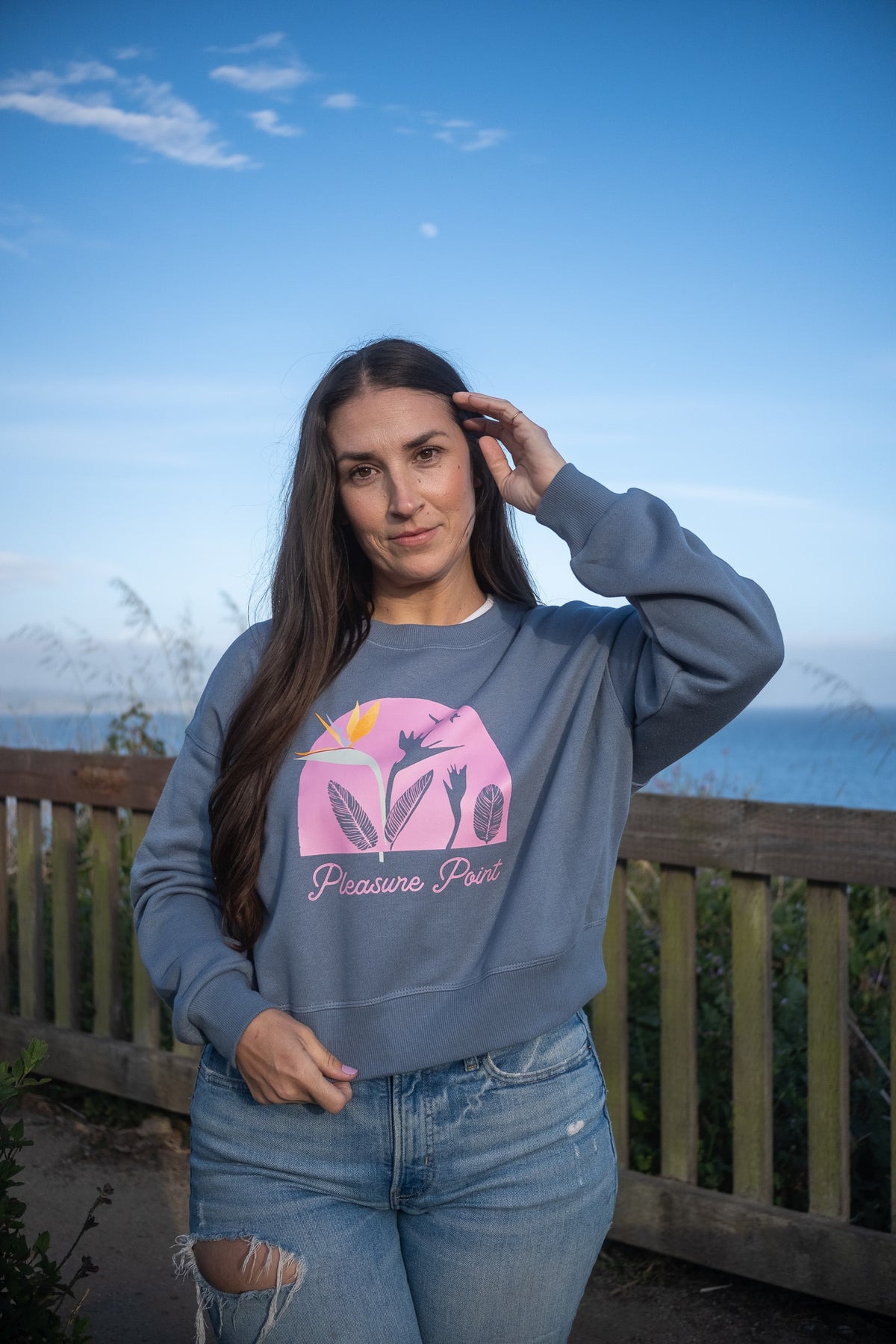 A woman wearing a slate blue crewneck sweatshirt with a pink graphic and the text 'Pleasure Port' stands outdoors in front of a wooden fence and blue sky.