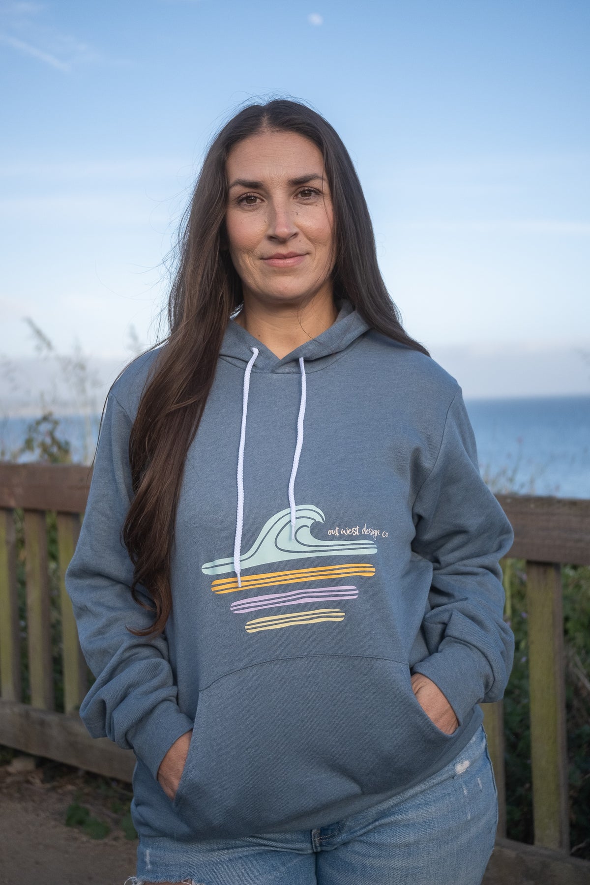 Woman wearing a blue hoodie with a retro wave design, standing outdoors with a scenic background.