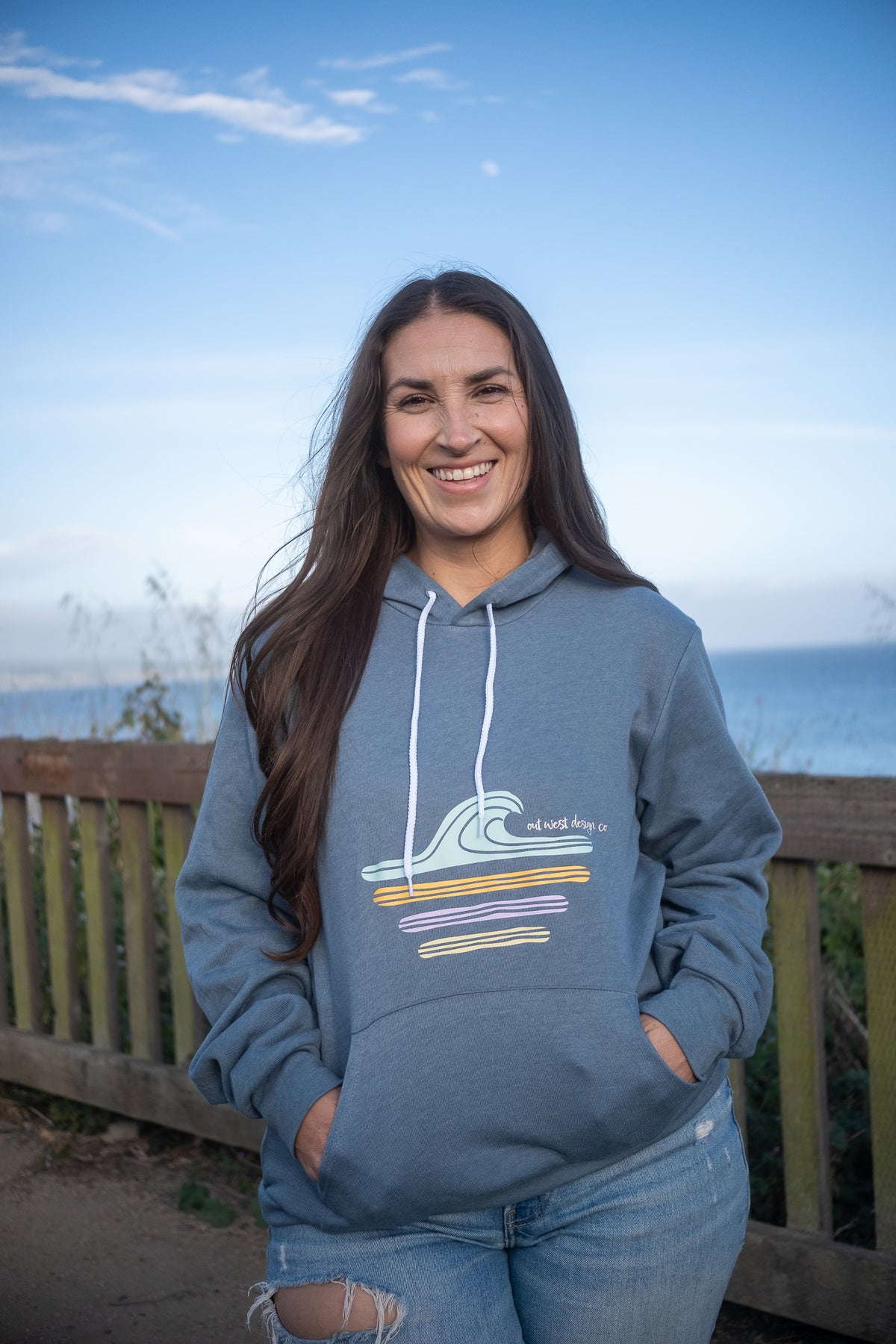 A woman smiling outdoors in front of a wooden railing and ocean view, wearing a slate blue hoodie with a multicolor wave graphic and white drawstrings.