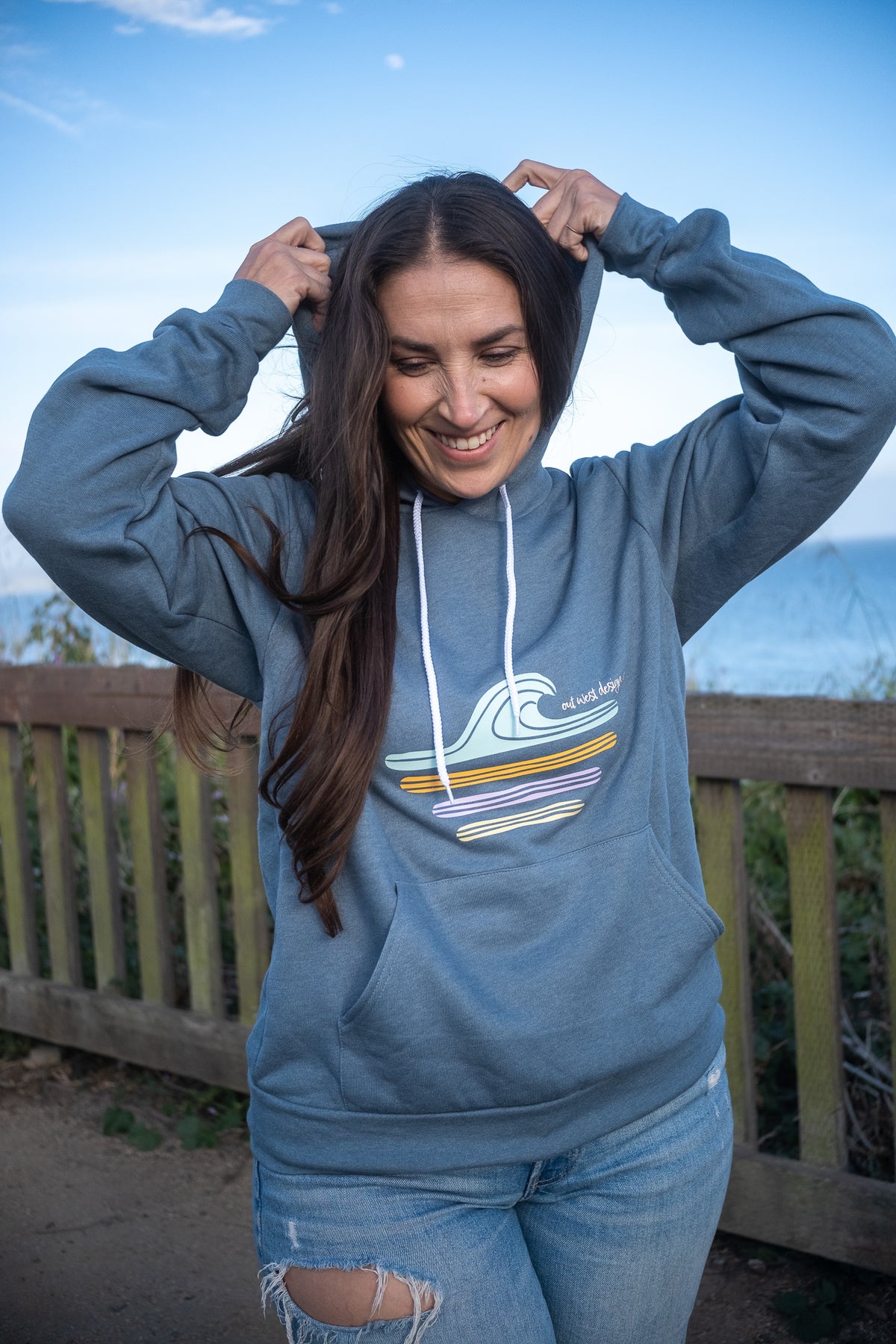 A woman wearing a slate blue hoodie with a multicolor wave graphic, posing outdoors in front of a wooden fence and ocean background.