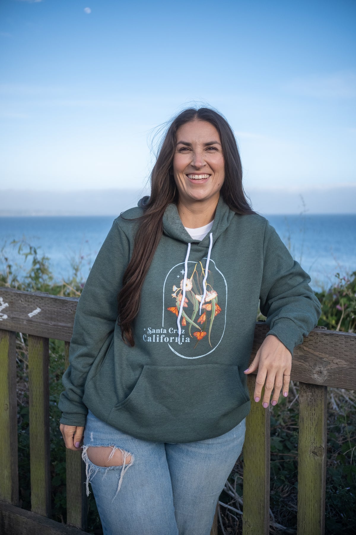 A woman smiling while wearing a dark green graphic hoodie with 'Santa Cruz California' and a monarch butterfly design, standing outdoors on a wooden boardwalk with the ocean and blue sky in the background.
