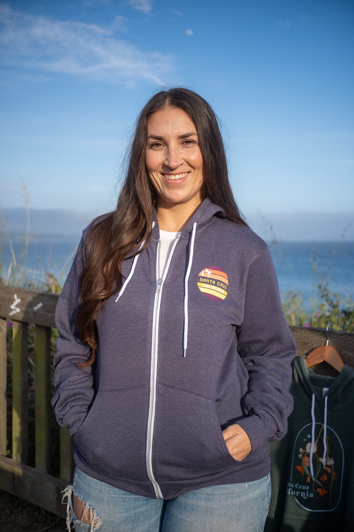 A smiling woman with long dark hair stands outdoors by a wooden railing with the ocean and blue sky behind her. She is wearing a dark slate purple zip-up hoodie with a small colorful chest graphic and white drawstrings.