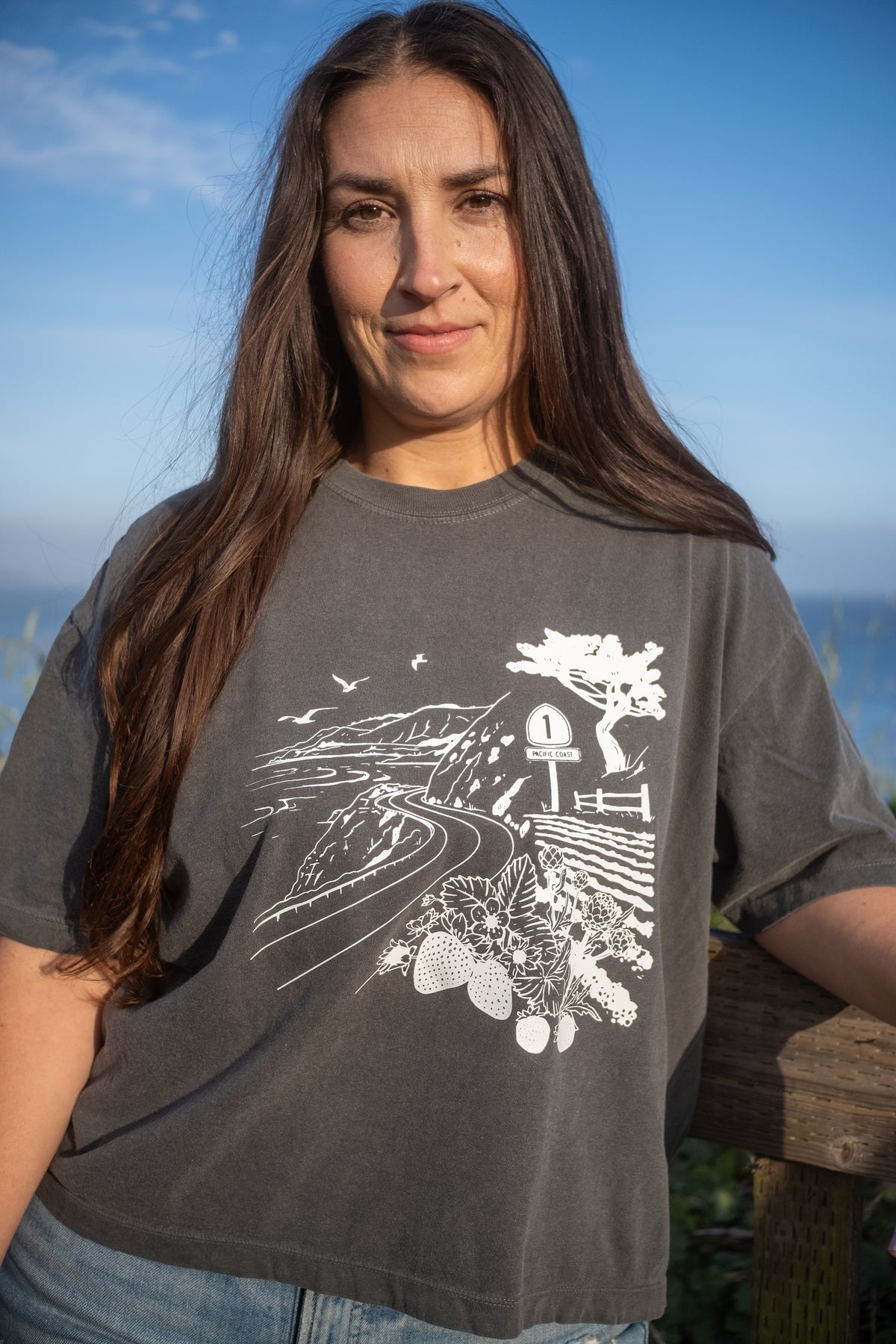 A woman with long dark hair wearing a dark gray graphic t-shirt with a white illustration of a hwy 1 landscape, standing outdoors against a blue sky and ocean background.