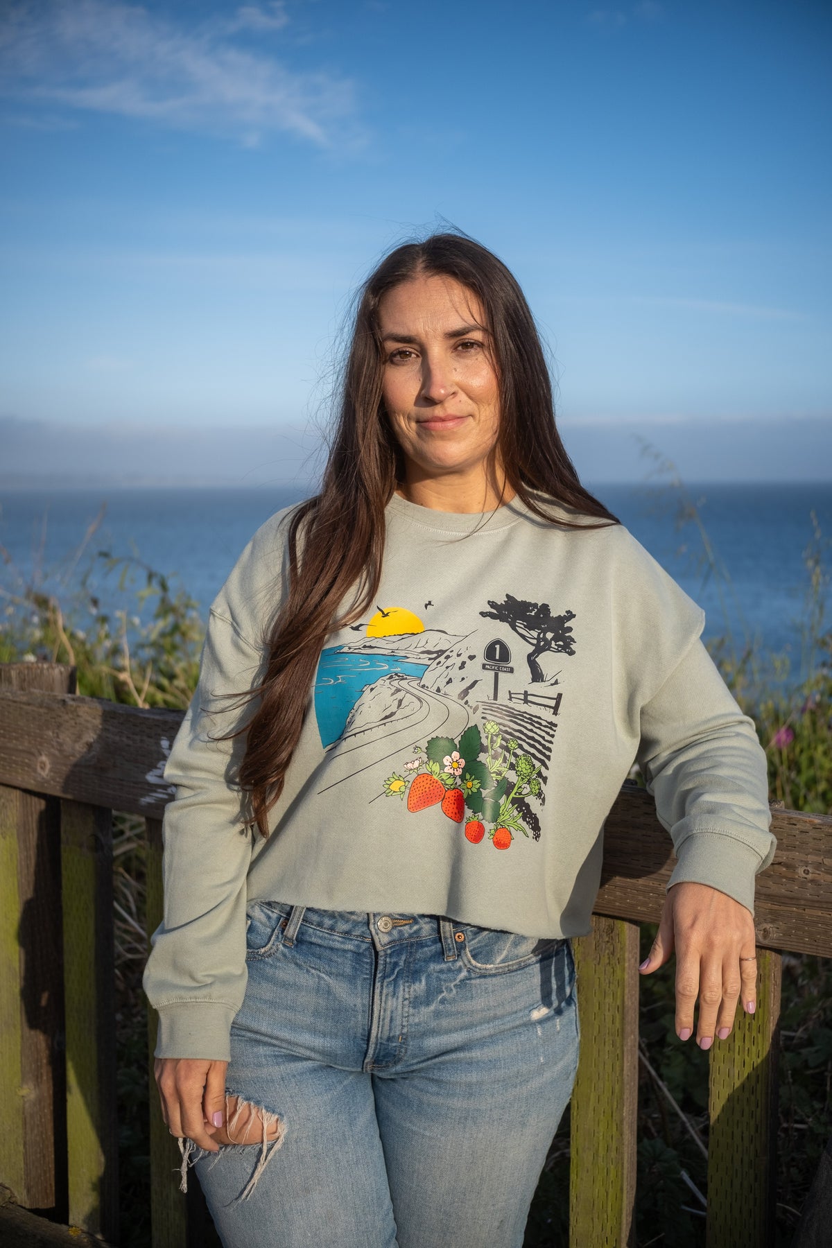 A woman wearing a light sage green graphic sweatshirt and blue jeans, standing outdoors on a wooden railing with a scenic ocean and blue sky background.