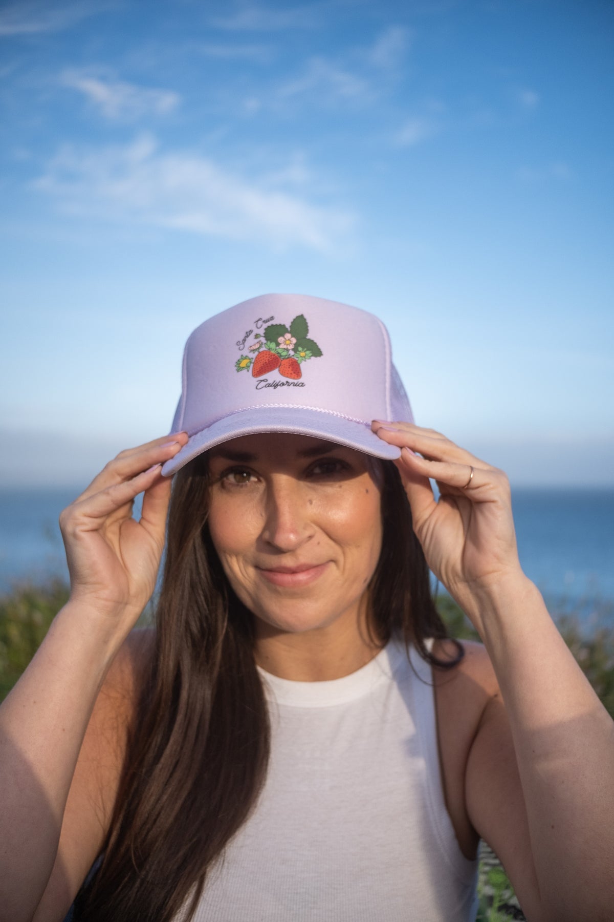 A woman outdoors by the ocean wearing a light lavender/purple trucker hat with a strawberry graphic and the brand name 'California' embroidered on the front. She is holding the brim of the hat with both hands.