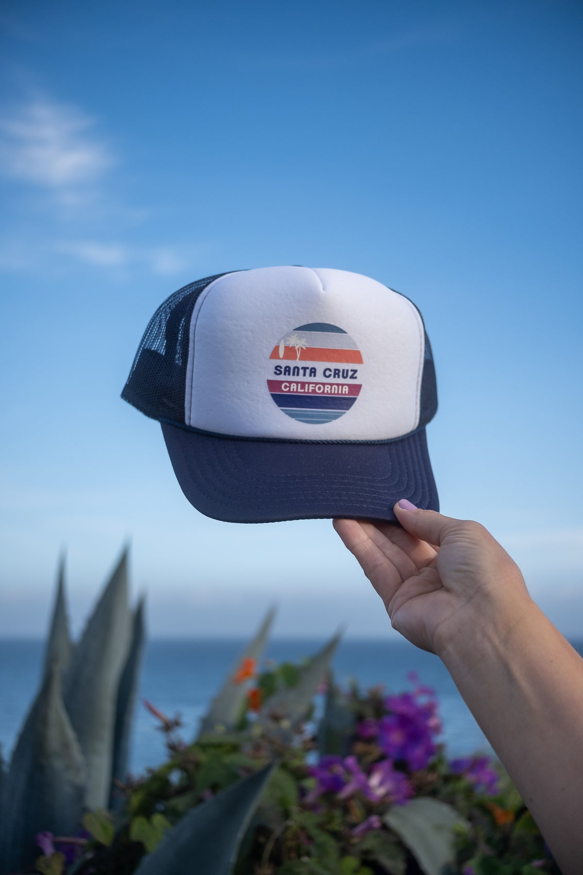 A hand holds up a white and navy blue trucker hat with a circular 'San Francisco California' graphic on the front, set against a blurred background of blue sky, ocean, and green foliage.