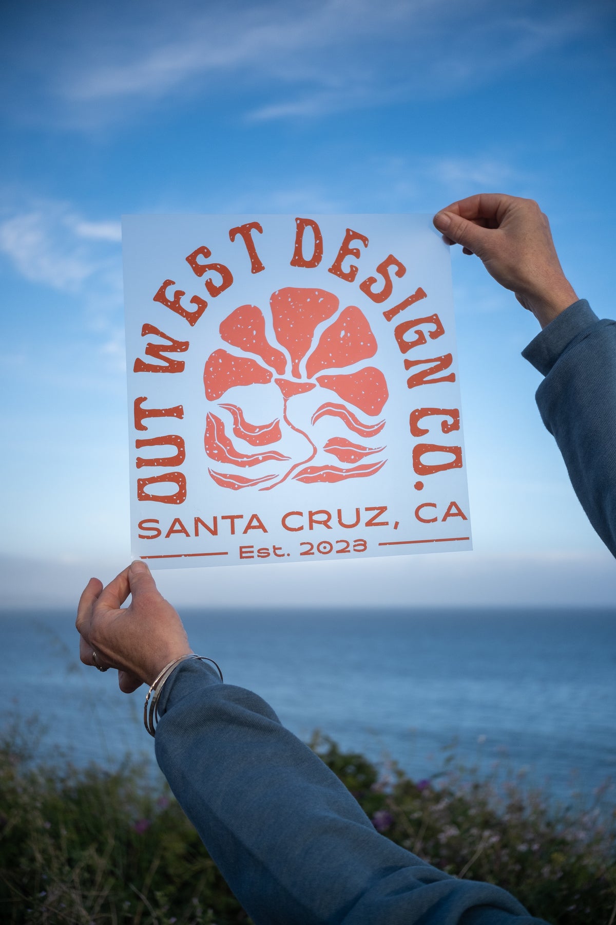 A person outdoors by the ocean holds up a white sticker or print featuring an orange coral reef logo and the text 'OUT WEST DESIGN CO. SANTA CRUZ, CA Est. 2023'