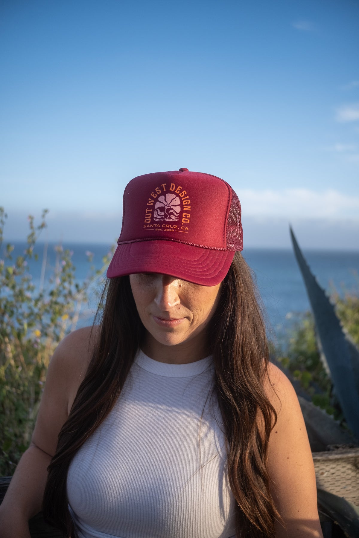 A woman with long brown hair wearing a light blue trucker hat with an orange circular logo, standing outdoors with the ocean and blue sky in the background.