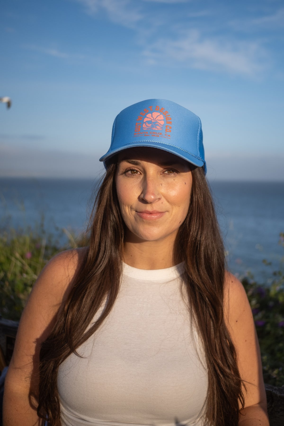 A woman with long brown hair wearing a light blue trucker hat with an orange circular logo, standing outdoors with the ocean and blue sky in the background.