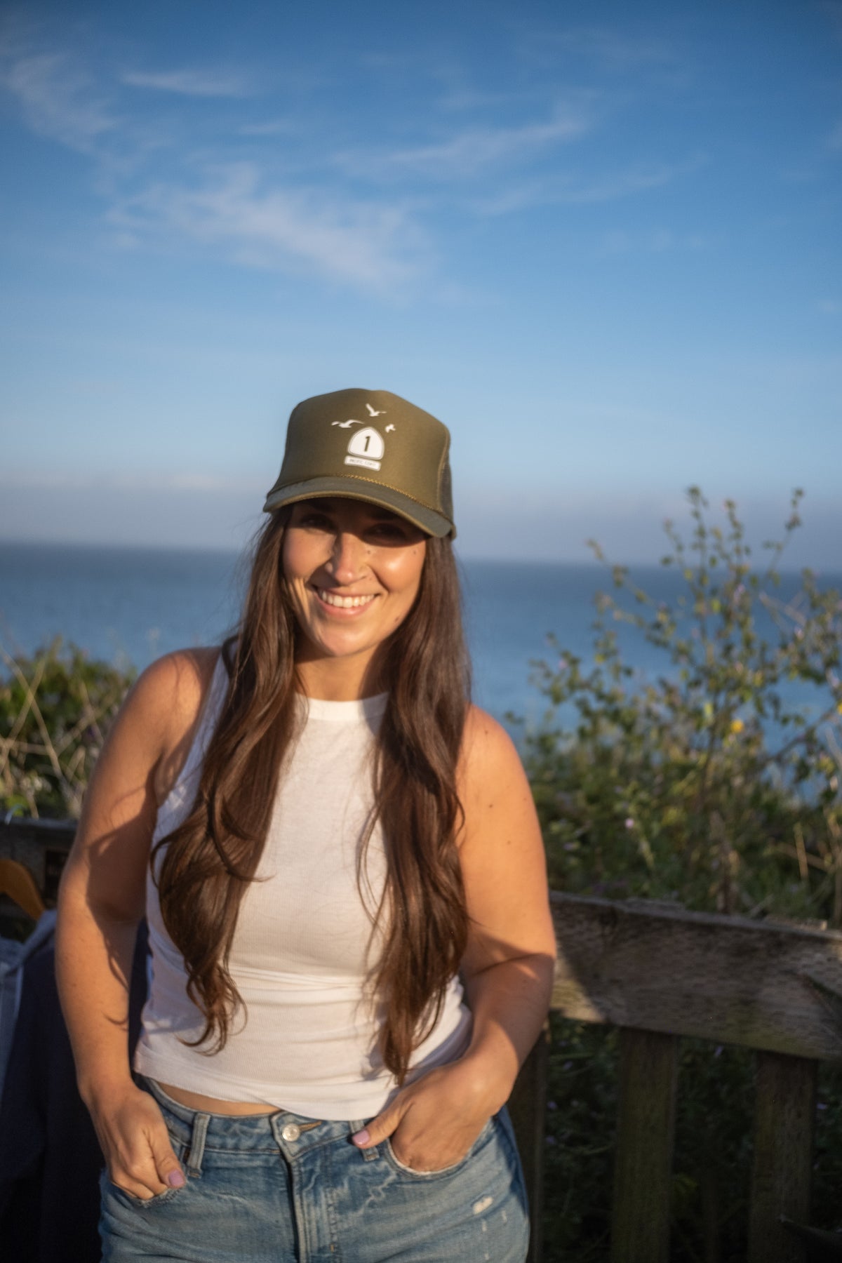 A smiling woman outdoors by the ocean at sunset, wearing an olive green trucker-style baseball cap with a white logo, a white tank top, and blue jeans.