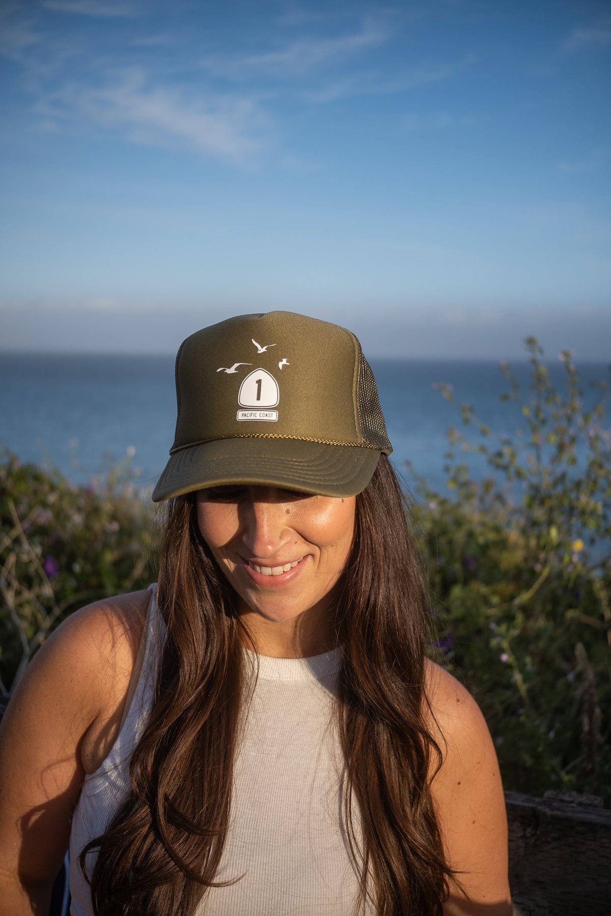A woman outdoors wearing an olive green trucker hat with a white shield logo featuring a bird and the number 1, with a blue ocean and sky in the background.